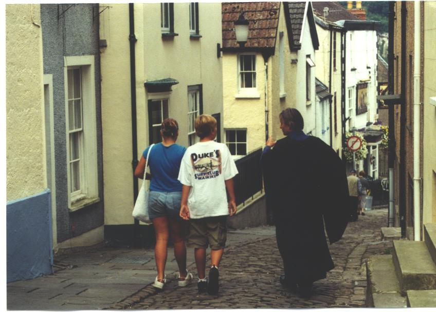 very steep, winding cobbled street with man in robes and two people in hiking gear walking away down it