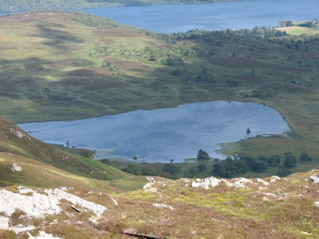 view down and out over small loch with large loch in the distance, with a low hill topped by scrubby trees in between