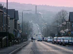 photograph looking down steep road, with factory chimneys and countryside beyond them