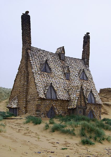 three-storey cottage covered in brown shells, standing on a beach, with very high, steeply-pitched roof with two rows of dormer windows, one above the other