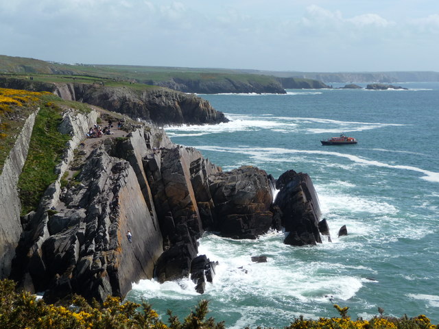 Sheer smooth black cliffs with a tiny human figure scaling them and a boat in the distance