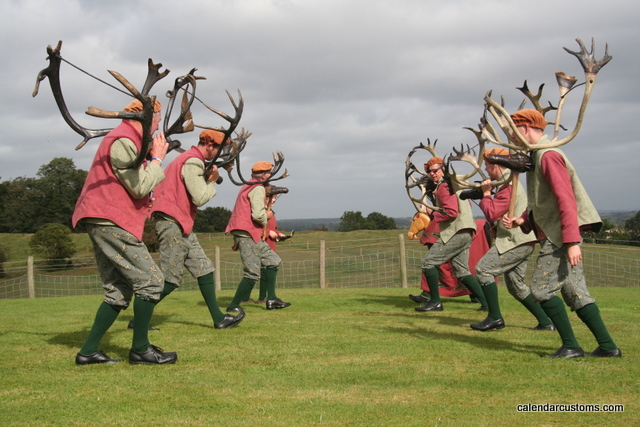 Photo of two rows of three men each in pink jackets, grey trousers and black boots, carrying antlered deer-skulls and dancing to each other