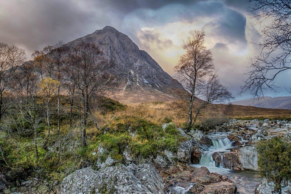 view of a high, dark, conical mountain, with in the foreground a stream tumbling among rocks and a few half-bare, scrubby trees