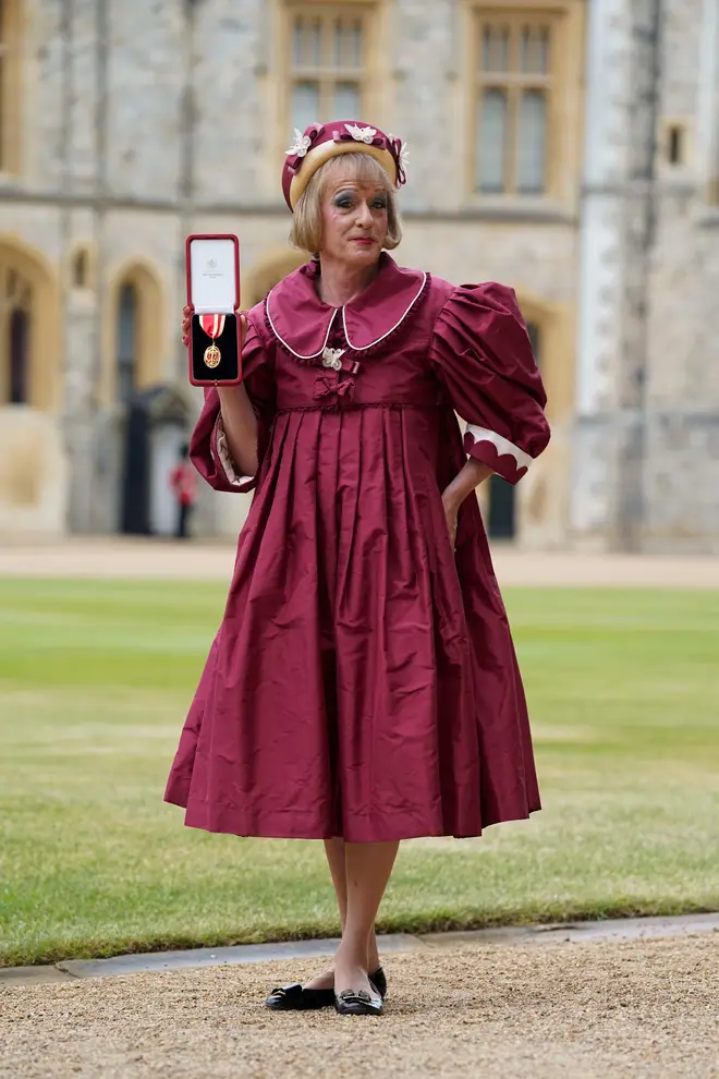 Photo of a tall blond man wearing a maroon dress and ladies\' hat, and holding up an award