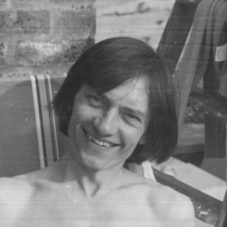 black and white head-and-shoulders shot of John lounging bare-chested on a deckchair, grinning at the viewer, with his head tilted to his left and long curtains of black hair flopping across his forhead