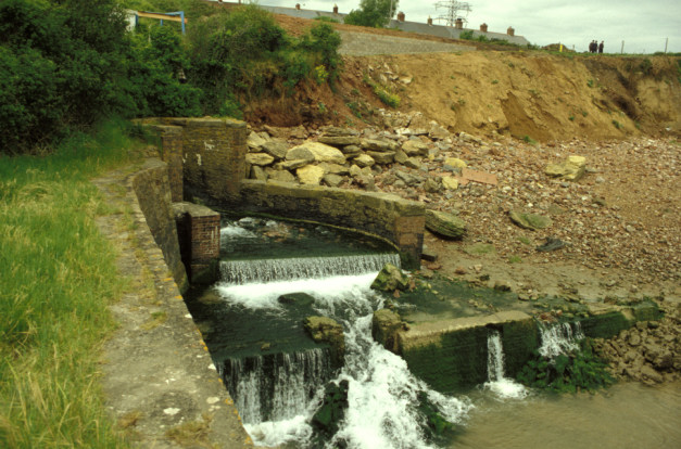 water pouring out from a brick culvert to form a small waterfall, at the foot of an earth bank with houses glimpsed at the top