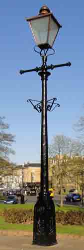 photo showing old-fashioned iron lamppost with glass cage-style top and crossbar, in front of a modern suburban scene