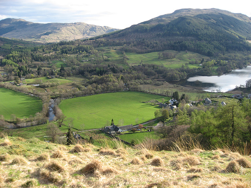 looking down from a high place into a valley full of green fields and the corner of a loch, with mountains rising in the distance