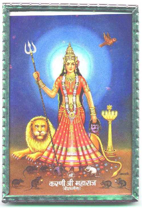 Hindu-style icon of crowned woman in red dress standing in front of a reclining lion, carrying a trident and surrounded by several brown rats and one white one, who are eating sweets off the floor by her feet
