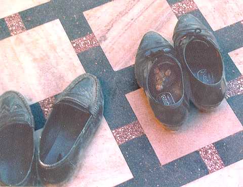 Two pairs of black shoes on a pink and grey marble floor, with a rat sitting in one of the shoes