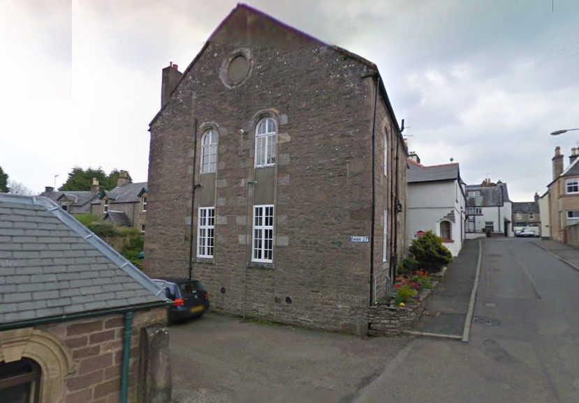 looking up a steep road lined with small white buildings, past a larger building in irregular brown stone with bushes in front of it, the end wall having two pairs of white-painted windows divided into small panes, the windows on the upper floor having arched tops