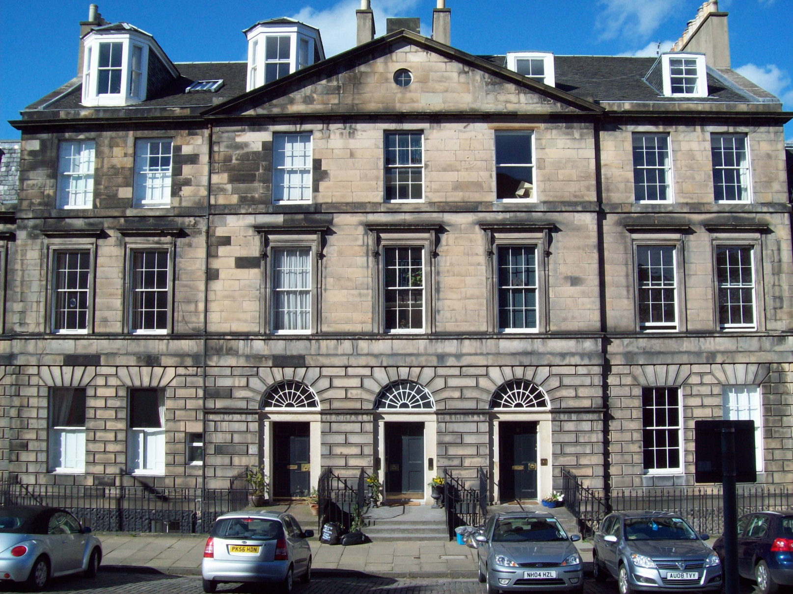 very large, grey-beige Georgian building having a central section with three doors, each accessed by shallow steps bridging a basement area, each with a fanlight over the door and then two storeys of tall windows, topped with a peaked roof with a dormer window either side of the peak; to either side of the central, peaked block is a block with a flatter roof having no doors but three storeys of windows, two abreast, plus two basement windows and a single dormer in the roof