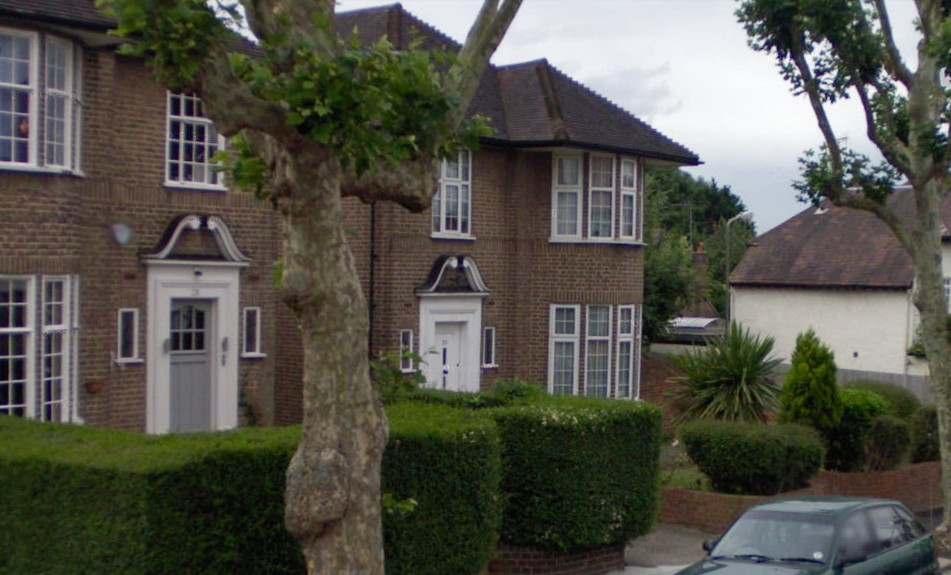 view looking across the front of a two-storey building in small brown brick towards another ditto, with the corner of a cream-painted house visible beyond: the brown buildings have rounded bays with tall white-framed windows divided into small panes, steep dark roofs and large front doors set in white surrounds with curly mouldings over the top; both brown houses have low brick walls around their front gardens and the nearer one also has a high squared-off privet hedge, and both have a gnarled tree growing out of the pavement in front of them