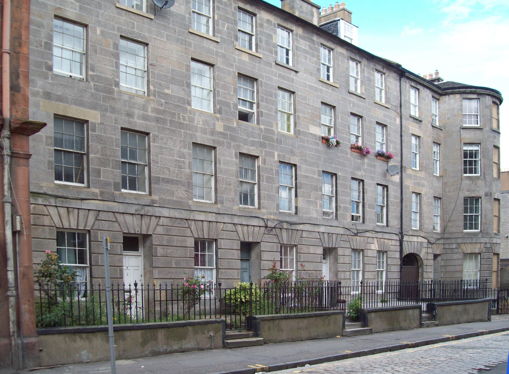 row of flat-fronted, four-storey buildings made of smooth grey stone, with large oblong windows divided into small panes, and small paved front gardens, the whole row terminating in a round tower of the same height