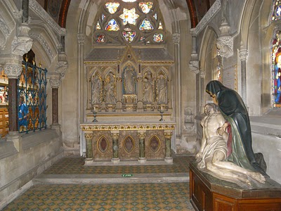 interior of Victorian Gothic church, looking towards an altar below a stone lacework window, with stained glass to the left and a polychrome pieta statue on the right