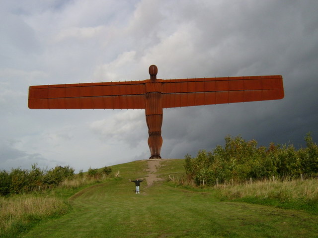 enormous, rust-red sculpture of a vaguely human figure with outstretched wings resembling those of an early plane, standing on a grassy mound and silhouetted against a lowering sky: the whole surface of the figure is divided into vertical ribs or vanes like those seen on the sort of Christmas decoration which unfurls from flat cardboard to a 3D shape