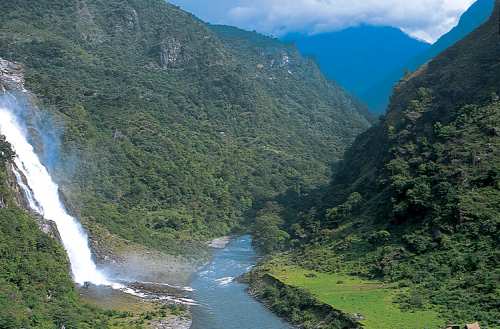 a steep-sided, wooded gorge with a river at the bottom, a white waterfall streaming down from the right and blue hills in the distance