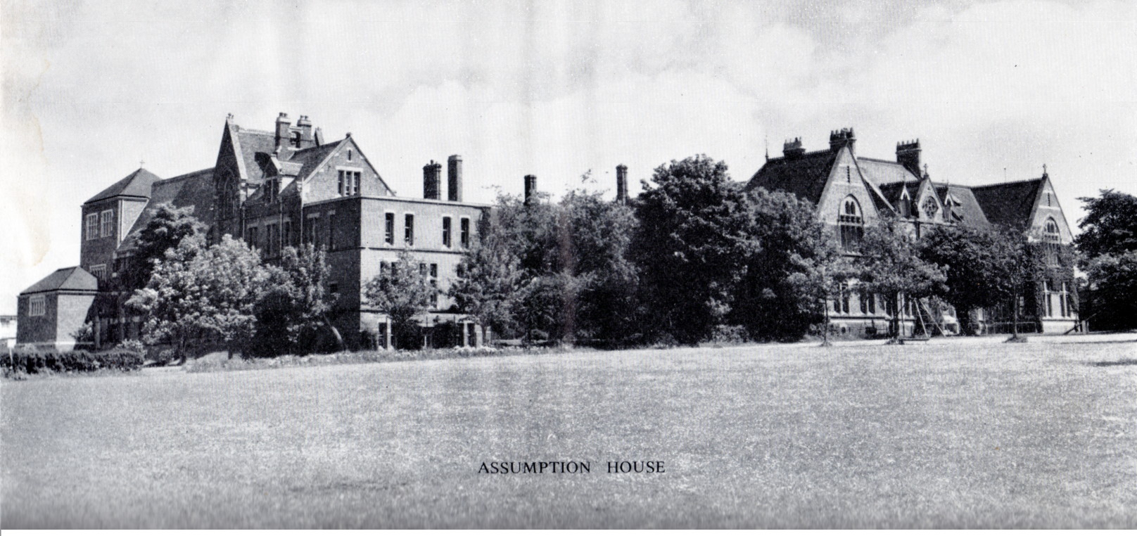 greyscale photo\' looking across a lawn to two large Victorian Gothic brick buildings fringed with trees