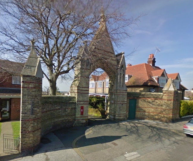 arched gateway decorated with religious statues and flanked by low, curved walls ending in stubby pillars, all in pale grey-brown stone banded with brick red