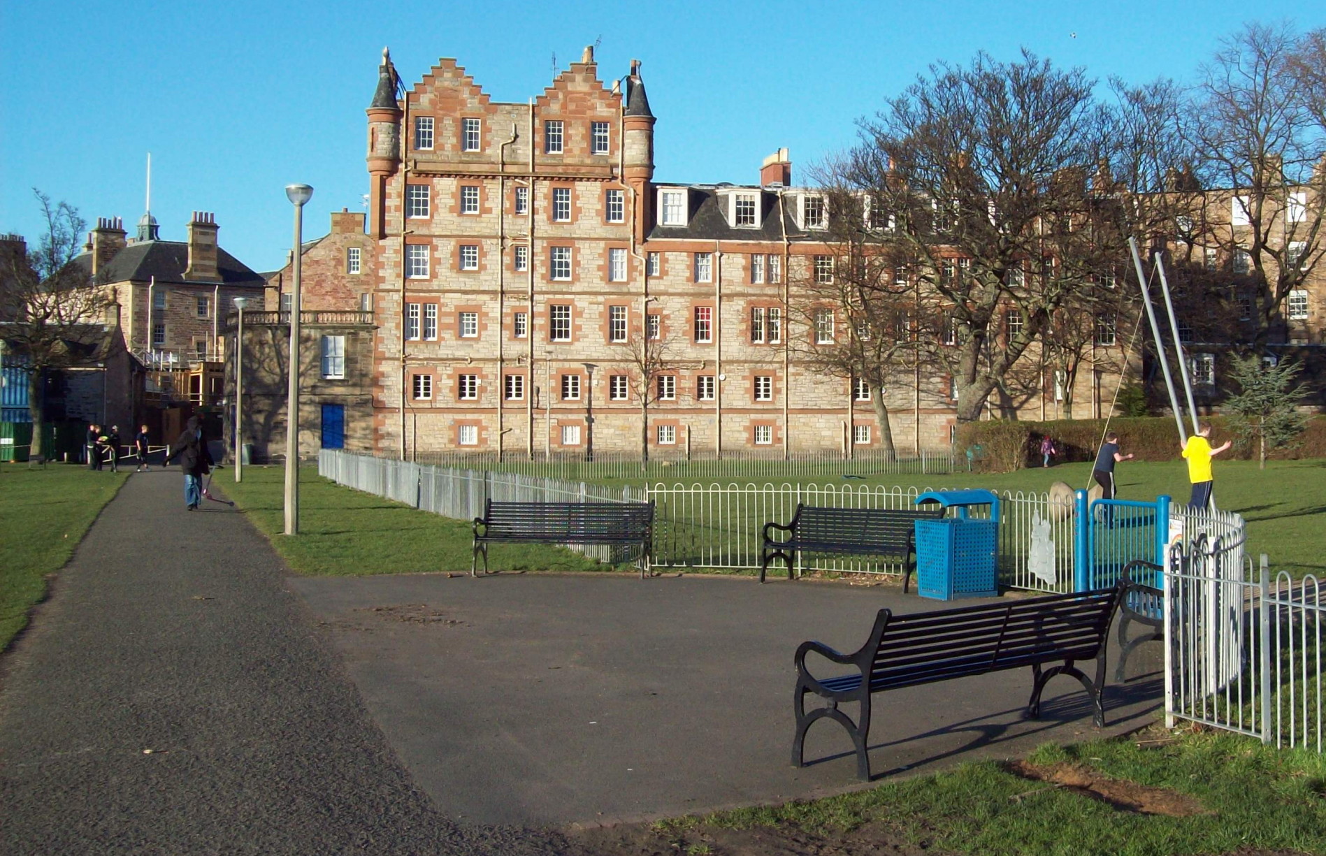 view along a concrete path across grass with a children\'s playground and cast-iron benches to the right of the path, and beyond the playground a flat-fronted strip of high buildings made of ginger stone: the left-hand end of the row ends in a higher six-storey section with a double triangular roof