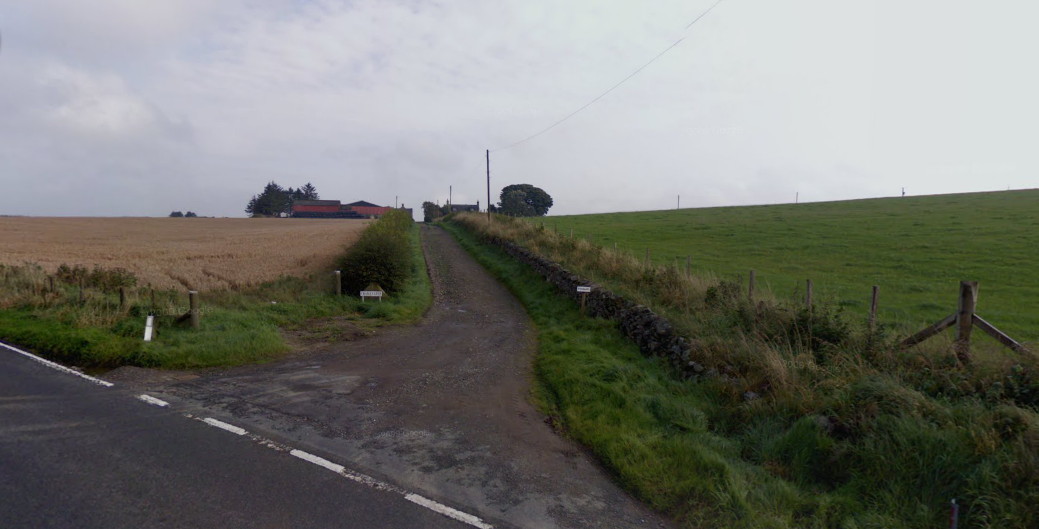 View along a narrow lane across fields to a cluster of farm buildings among trees