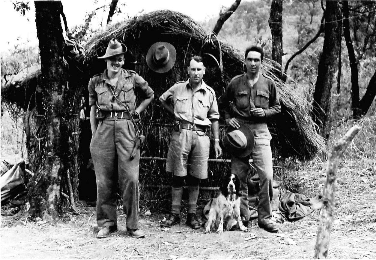 greyscale photo\' showing three men in casual military gear standing in a row in front of a rough straw shack, with a black and white spaniel sitting in front of them