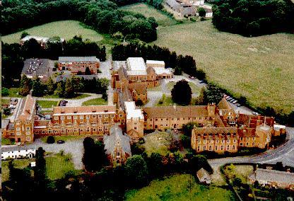 aerial view of a large complex of long Victorian buildings in ginger brick with cream trim, set amongst lush green fields and ornamental hedges