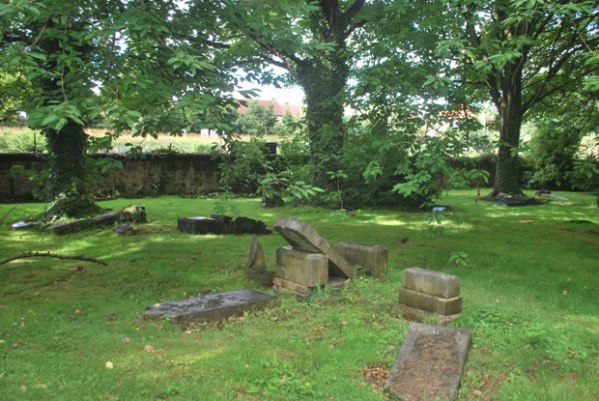 old, tumbled gravestones in a brilliant green wood, with a low wall and a sunlit field beyond it visible through the trees