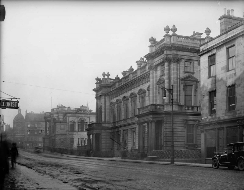 black and white photo\' of a glancing view across a street to a long three-storey stone building, the lower two floors being very high, with a complex parapet along the top and at either end a large, pillared porch