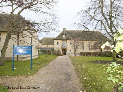 view along a concrete path across a grassy space to a large, Art Deco-ish two-storey cream building with a steep grey roof; part of a similar building is glimped to the left and in the near foreground is a free-standing blue sign with white lettering, announcing the Bate Collection of Musical Instruments