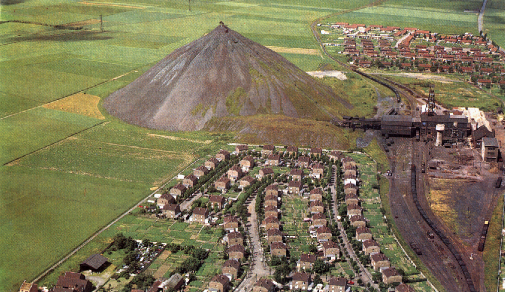 digital art showing a huge slag heap in a green field, with mine buildings to the right of it and streets of small houses also on the right