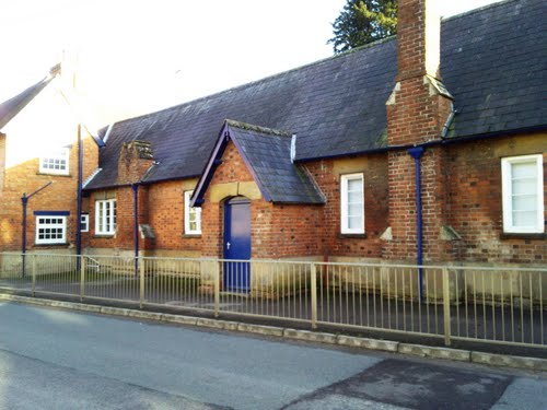 long low single-storey red-brick Victorian building fronting onto a narrow pavement edged with railing, and having a steeply-pitched dark roof, white windows, a porch with a blue door and two large chimneys, one of them broken off to a stub, which create bulges in the front of the building