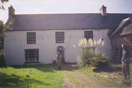 long, white-painted two-storey 18th C stone building, with a brown stone extension coming off it at right angles to the right of the picture, and a clump of pampas grass in the front garden