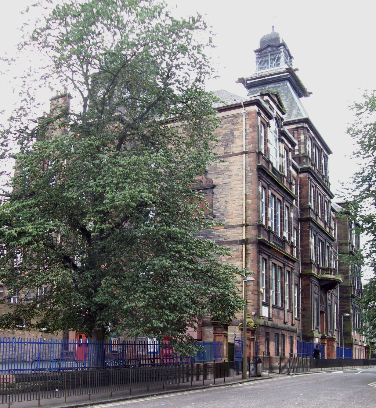 four-storey brownstone building with a clock-tower, and with a playground and ornate stone gates at its foot, in the foreground