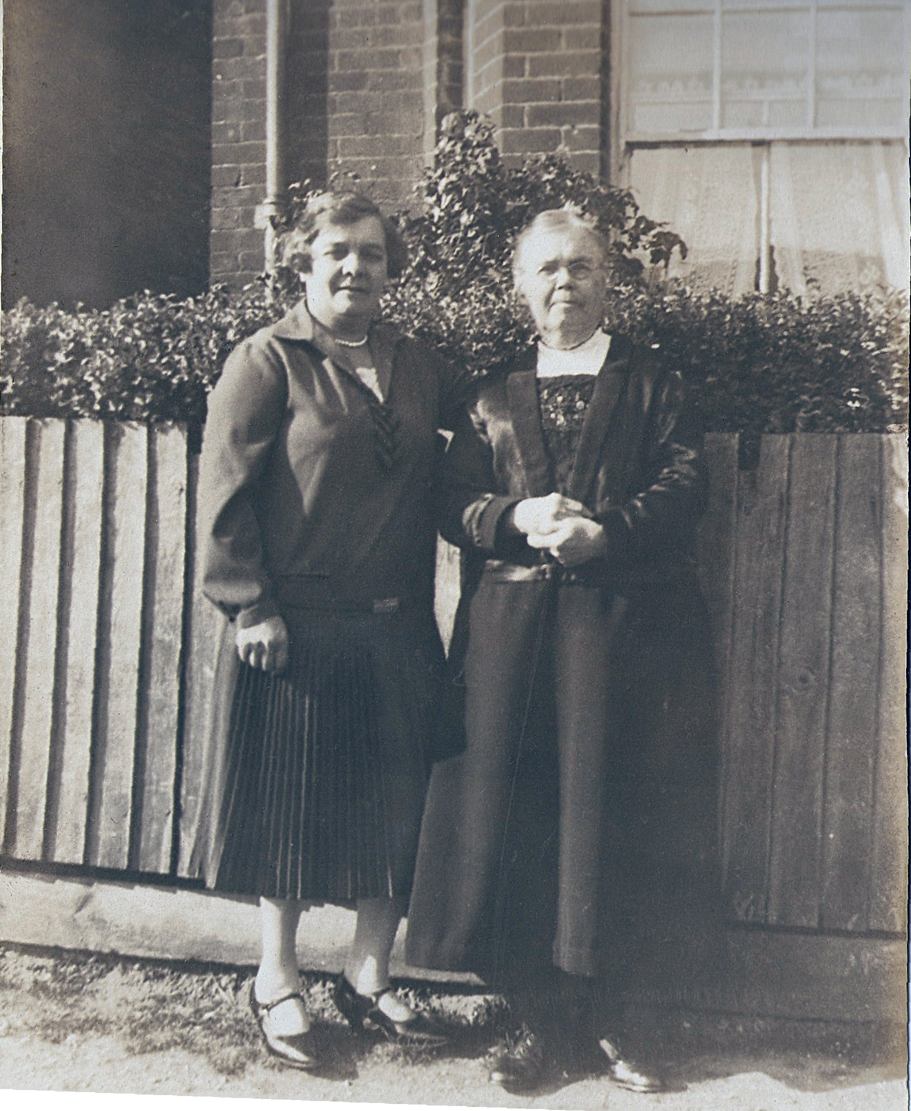 black and white photo\' of a middle-aged woman in a dark pullover and pleated skirt and an elderly woman in wire-rimmed spectacles and wearing a long black skirt and a black satin jacket, standing side-by-side by a fence in front of a house