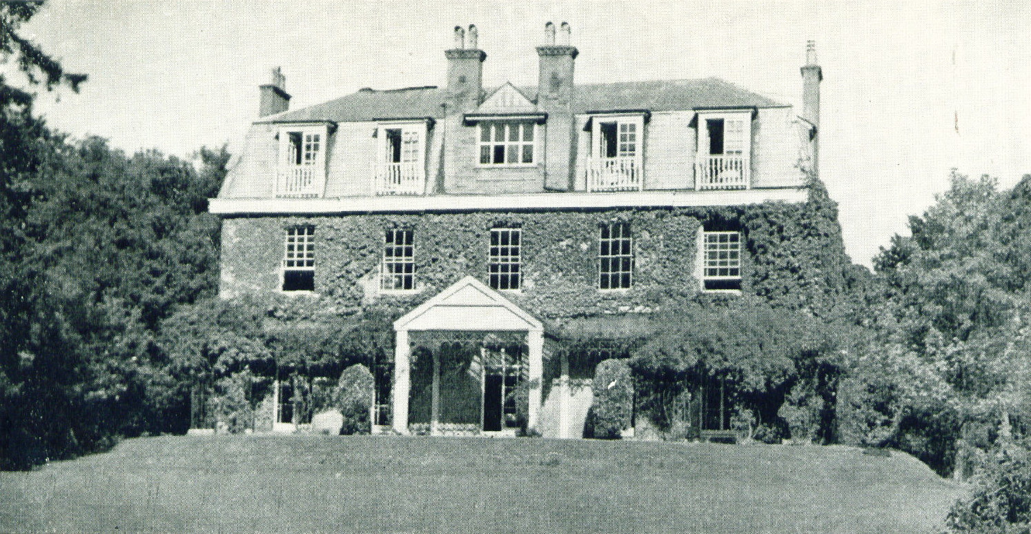 greyscale photo\' of a three-storey house dating from about 1820, having a very large porch flanked by a verandah on either side, a large central dormer window in the roof above the porch and two smaller dormers each side, elaborate chimneys, large windows divided into tiny panes, ivy growing over the walls and trees and bushes behind and to either side