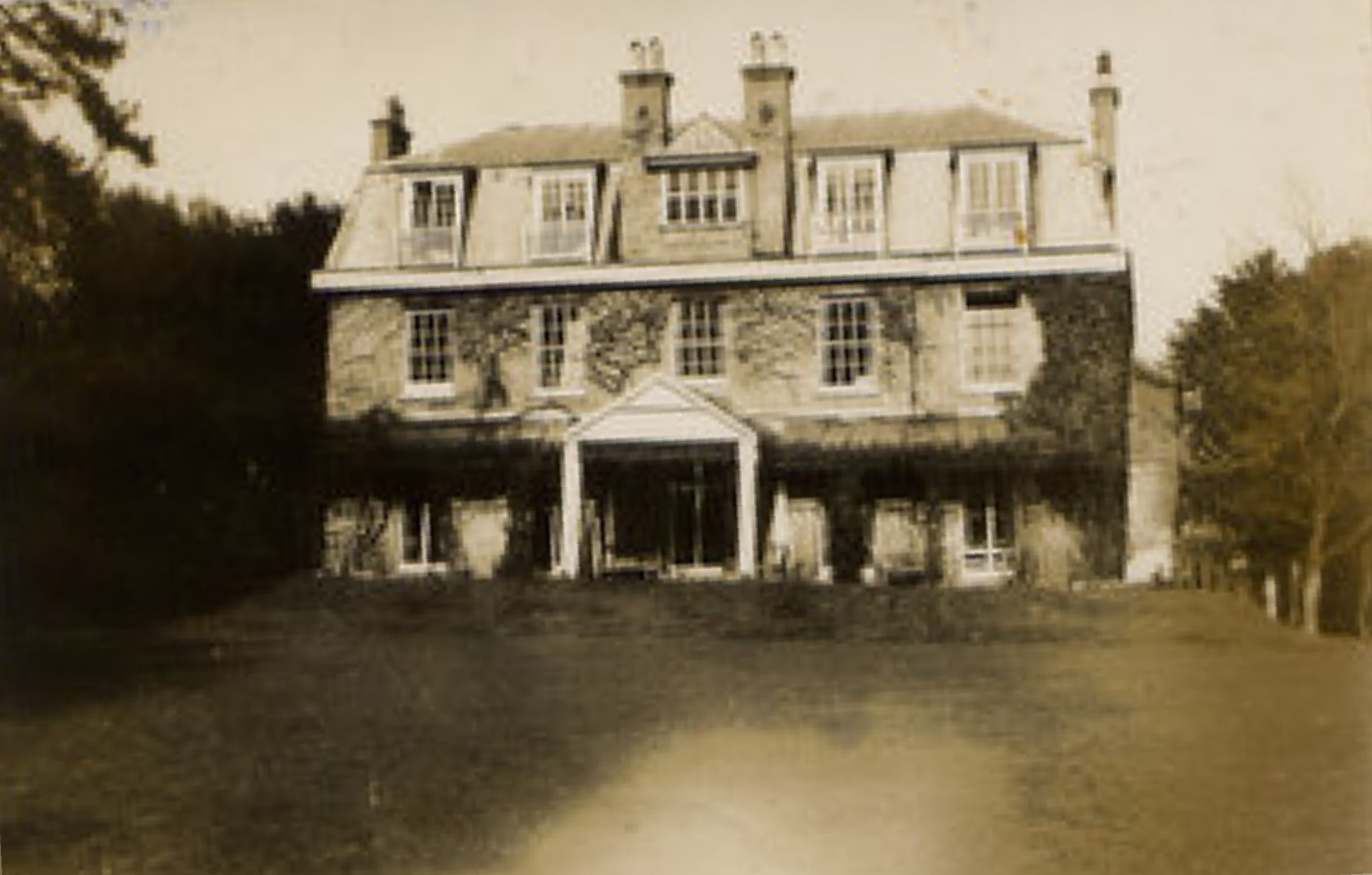 slightly fuzzy sepia photo\' of a three-storey house dating from about 1820, having a very large porch flanked by a verandah on either side, a large central dormer window in the roof above the porch and two smaller dormers each side, elaborate chimneys, large windows divided into tiny panes, ivy growing over the walls and trees and bushes behind and to either side