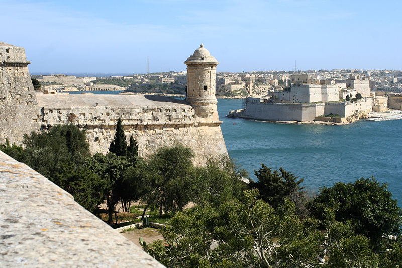 part of a pale, eroded fortress, with long low walls ending a little turret, on a promontory above a bay with a city visible on the far side