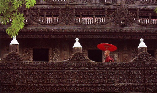 two layers of ornate dark-brown wooden walkways, the upper one with curved swags of open railings, the lower with a solid barrier, pillars crown with white objects resembling the teat on a baby\'s bottle, and a young man in red robes and carrying a red parasol, visible from the upper chest and above