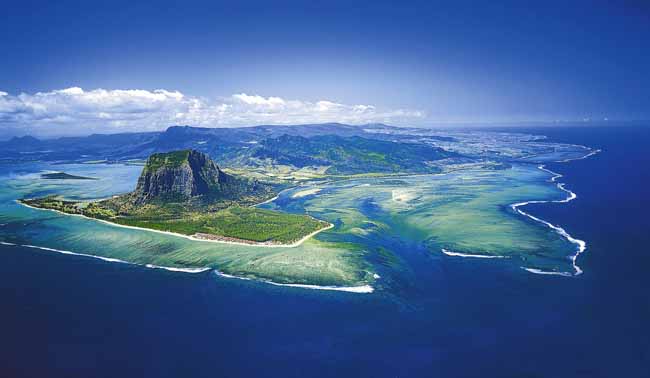 aerial photograph looking down on a roughly L-shaped island, with two blocks of low-lying, bright-green land, and a large flat-topped mountain where they intersect, set among deep-blue lagoons