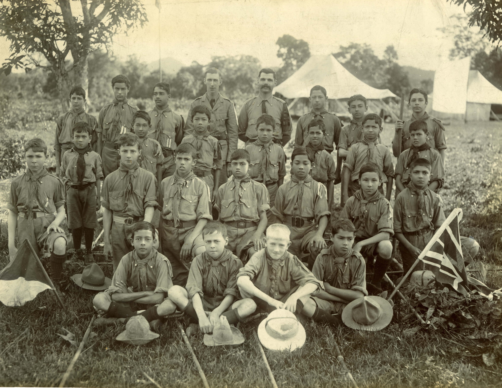 greyscale photo\' showing twenty-four boys and two young men, all in scout uniform, bareheaded, lined up in four rows with white tents behind them and flags displayed on either side