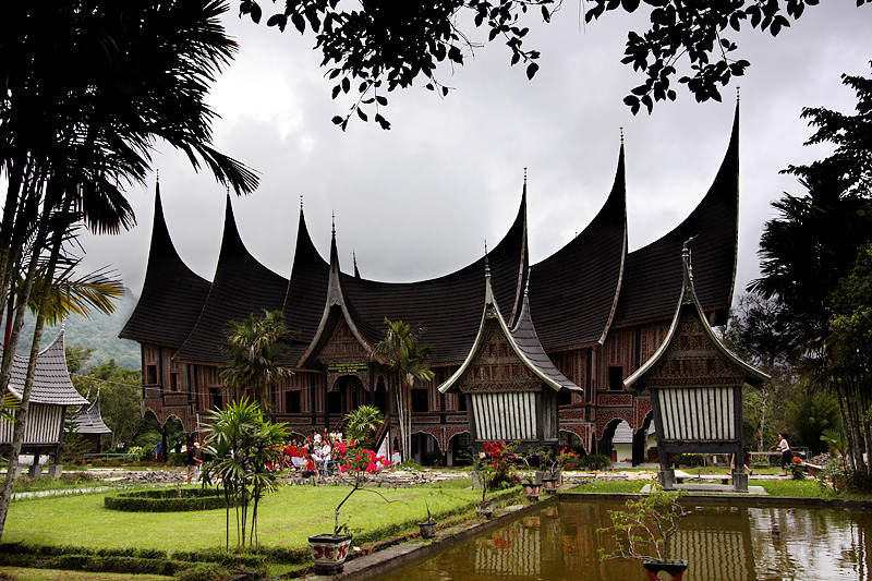 long two-storey building with smaller sections branching off at right-angles, coloured chocolate-brown divided into panels delineated by black and white lines, the whole built on short stilts which allow a series of small arched tunnels to run under the building, with end-walls which incline forwards and deeply-curved black roofs rising to sharp points at the gable-ends, with a garden and a stone-rimmed square pool in front of the building