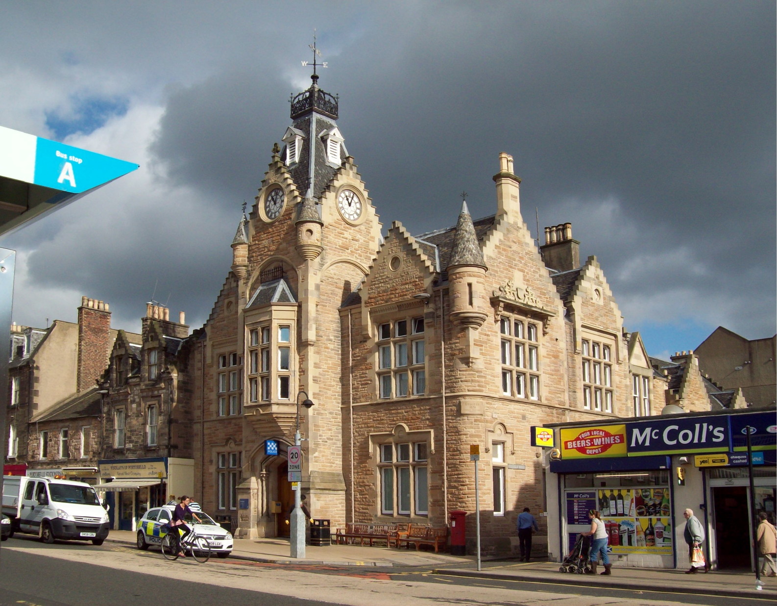 corner-on view of a large beige stone building crowned with elaborate turrets and pinked edges and a clock of which two faces are visible, resembling eyes, with storm-clouds gathering behind it