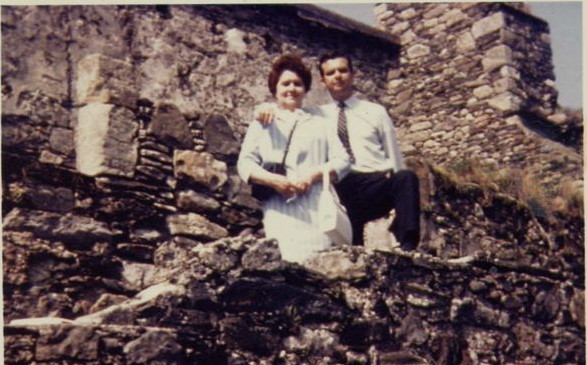 colour photo\' of a middle-aged woman in a white dress and a younger man in a white shirt and dark trousers, standing high up among the grey stones of a semi-ruined wall