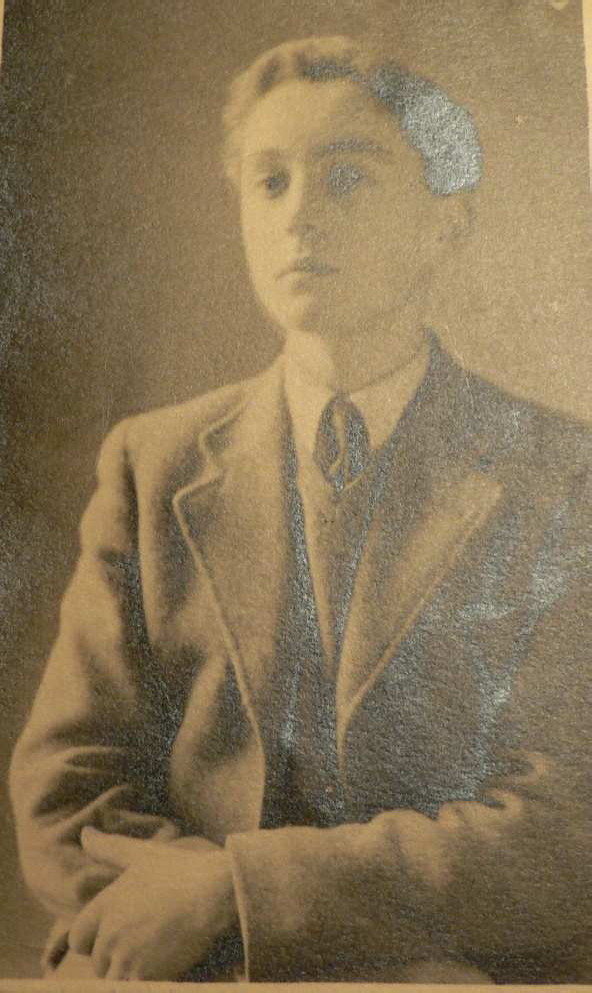 black and white upper-body portrait photo\' of calm, slightly snooty-looking boy aged about sixteen with wavy brown hair, wearing a lightish-coloured blazer, V-necked sweater, shirt and tie, with his arms loosely folded at waist height