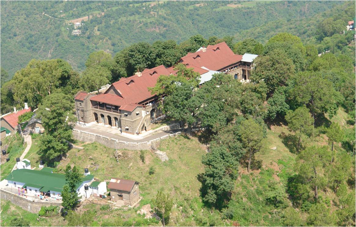 looking down on a large red-roofed buildingwhich is sited on a hill-top, with wooded land falling away behind it and rising again in the distance