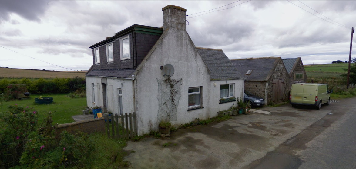 Small white farm cottage with two brown stone barns beyond it and a green garden on the far side, against a lowering sky