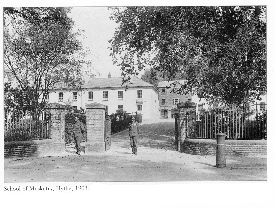 long two-storey white building in the middle ground, with a three-storey brown building set back behind it to the right; in front are gardens, a low wall with metal railings and a double gateway, one narrow for pedestrians, one wide for vehicles, marked out by three brick pillars and with two sentries standing in front