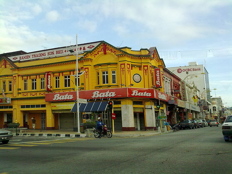 looking across a road towards an Art Deco-ish corner-block of shops painted mustard yellow with red trim, with a row of similar, slightly ramshackle shops disappearing into the middle distance to the right