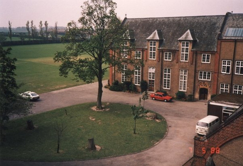 angle shot across a large two-storey red-brick building with a lawn, trees and a driveway in front of it